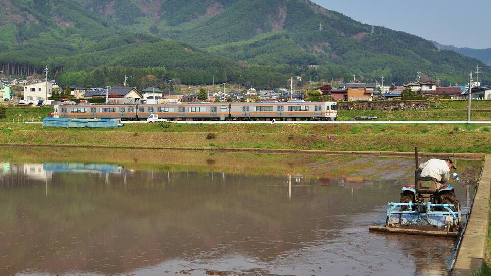 A train on tracks, a person on a tractor, and a mountainous landscape in the background.