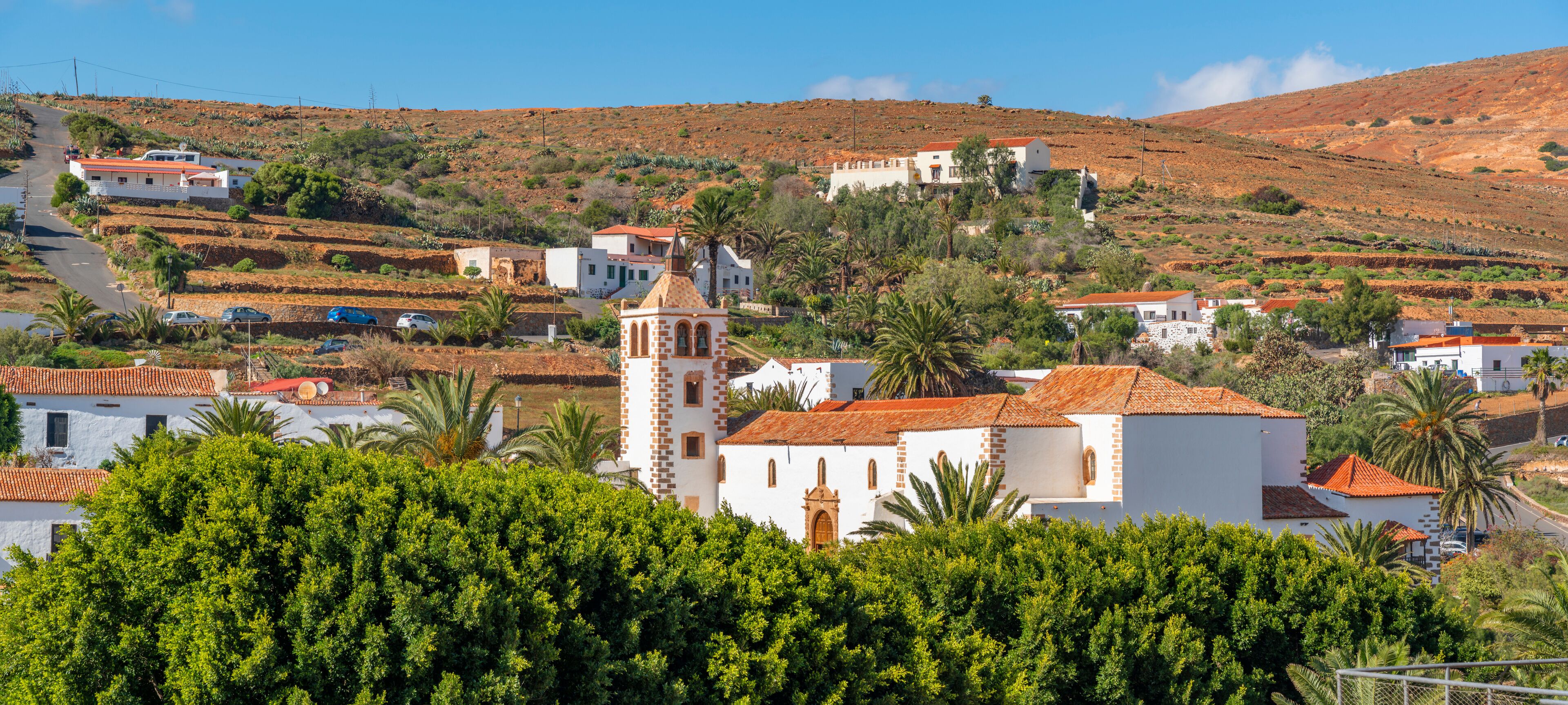 Uma vila situada numa encosta, com campos em socalcos, edifícios brancos com telhados vermelhos e uma torre de igreja imponente.