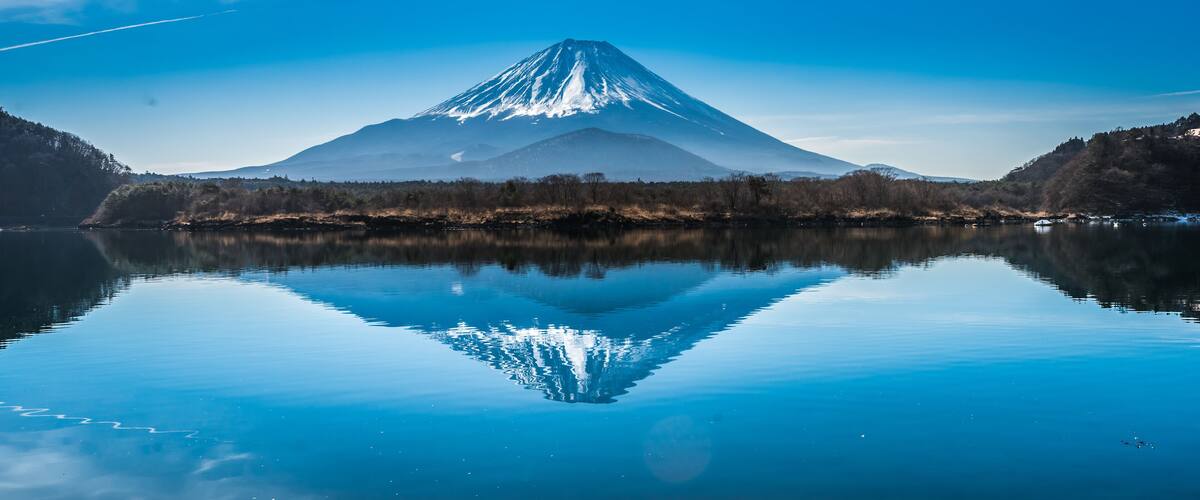 A snow-capped mountain reflected in a calm lake, surrounded by forested hills.