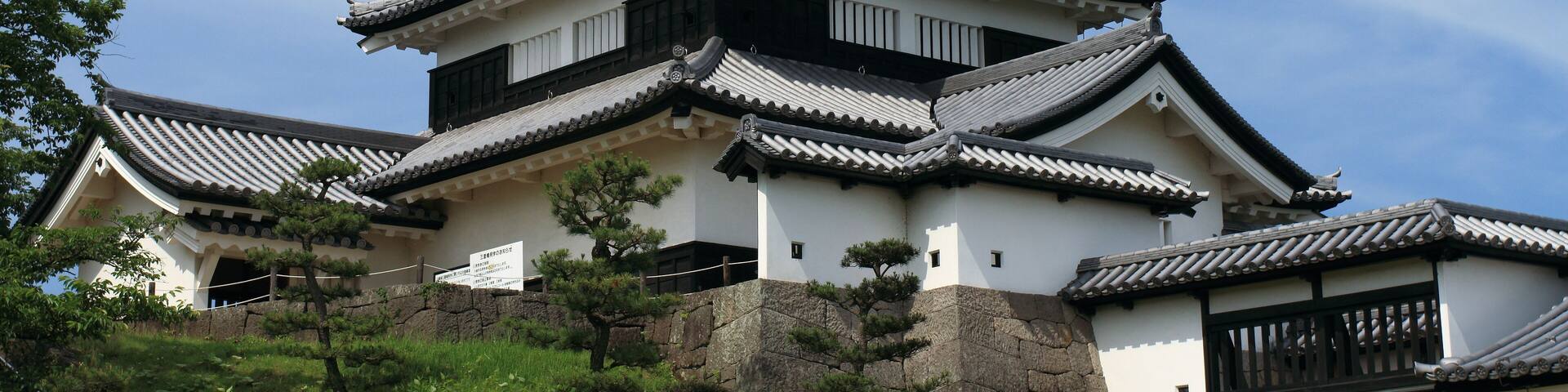 A traditional Japanese castle with multiple tiers, white walls, and black accents, surrounded by greenery and set against a clear sky.