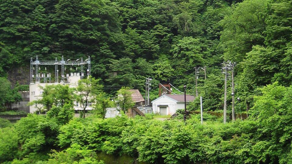 A river with a rocky outcrop in the foreground, surrounded by dense green forest and a hill with a tall structure.