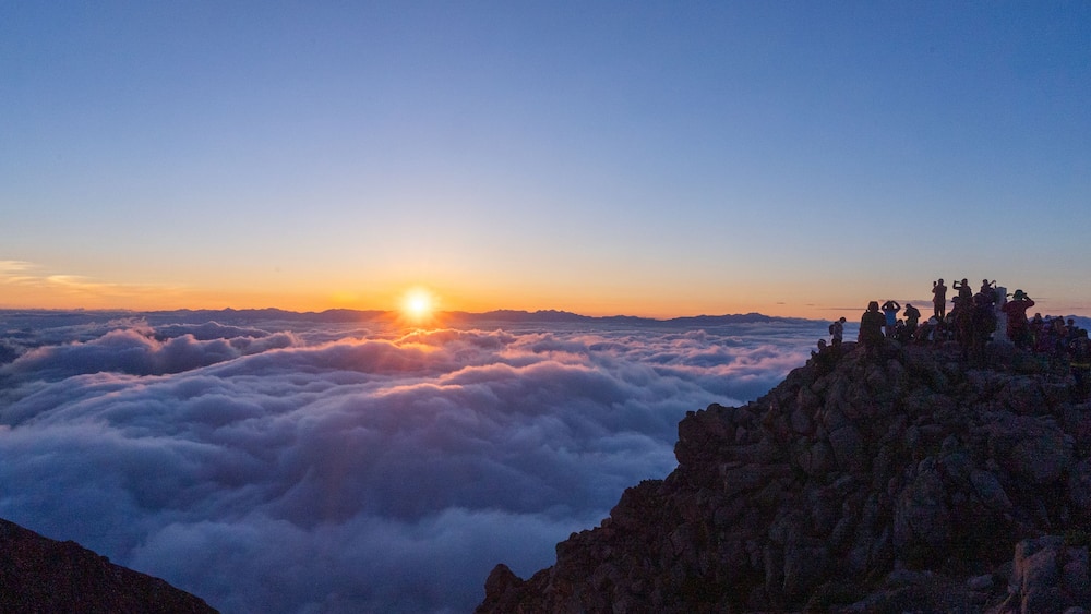 Sonnenaufgang über einem Gebirge mit Wolken darunter und Personen auf einem Felsen.