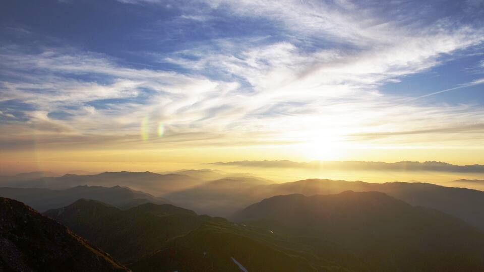 A mountain landscape with a sunrise or sunset, casting a warm glow over the peaks and valleys.