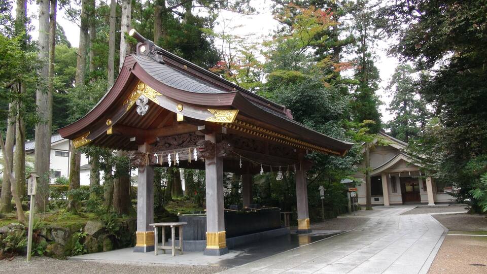 A traditional Japanese pavilion with ornate roof and golden accents, surrounded by lush greenery and tall trees.