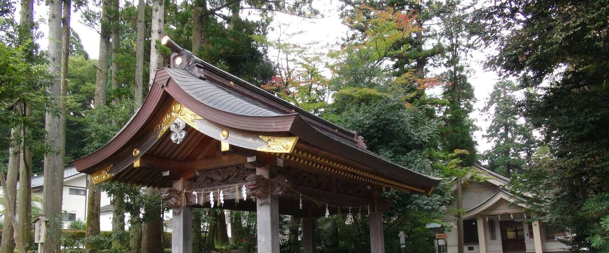 A traditional Japanese pavilion with ornate roof and golden accents, surrounded by lush greenery and tall trees.