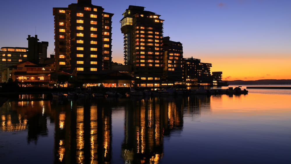 Ein Stadtbild am Wasser im Abenddämmerung, mit beleuchteten Gebäuden, die sich im ruhigen Wasser spiegeln.
