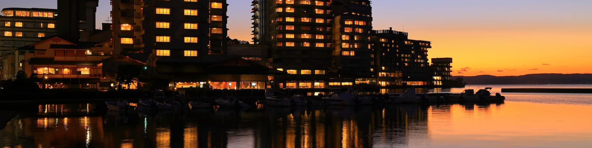 A waterfront cityscape at dusk with illuminated buildings reflecting on the calm water.