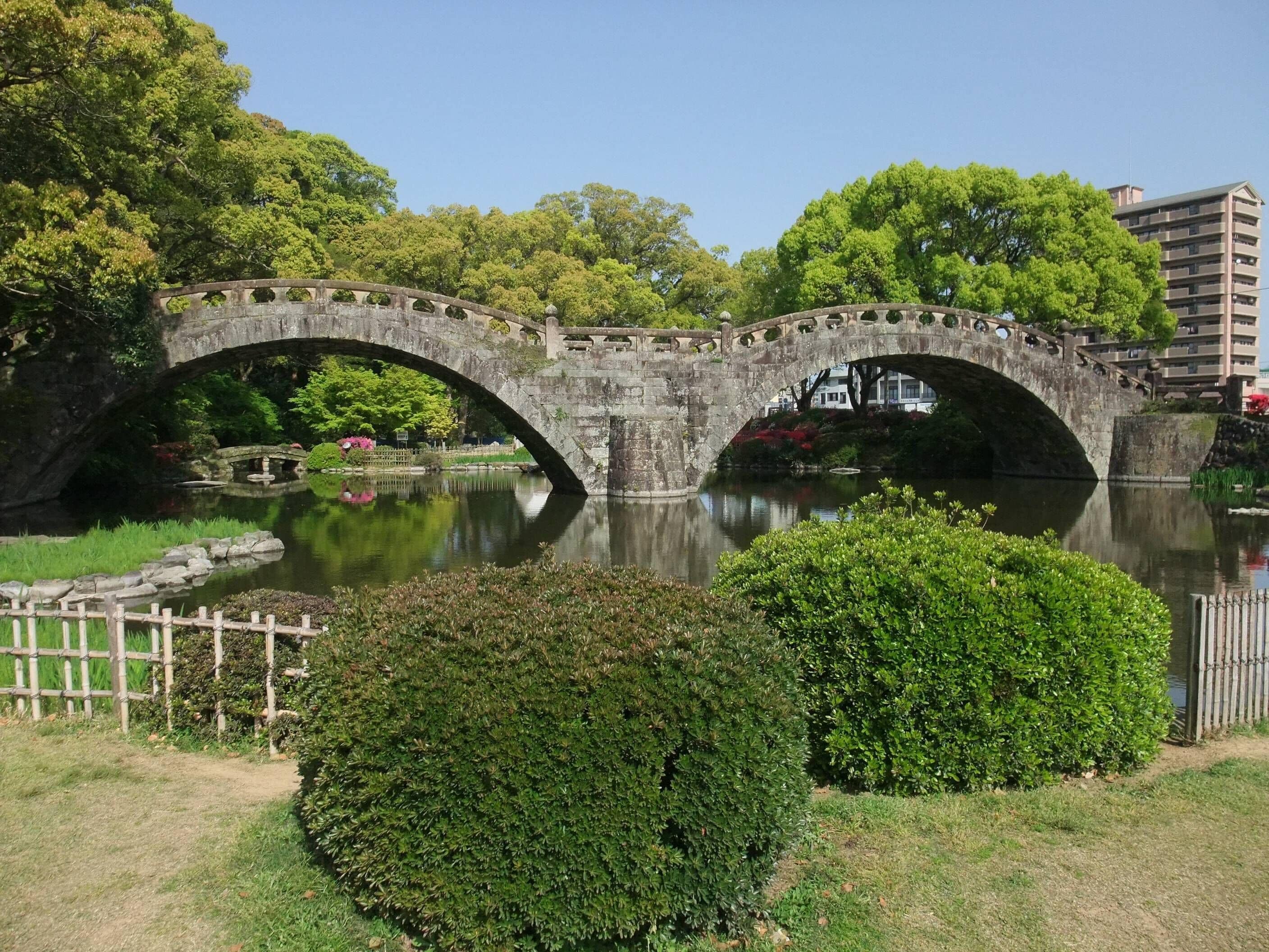 A stone bridge over a calm river, surrounded by lush greenery and trees.