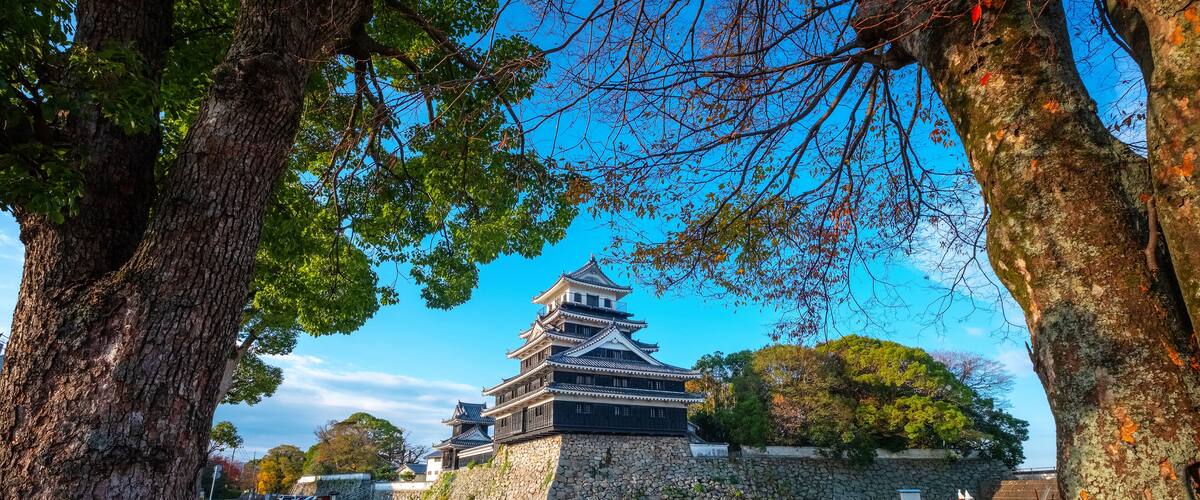 A traditional Japanese castle with multiple tiers, surrounded by trees and a clear blue sky.