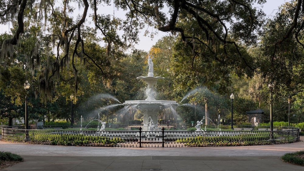 Une fontaine avec des jets d’eau, entourée d’une clôture, située dans un parc arboré sous un ciel dégagé.