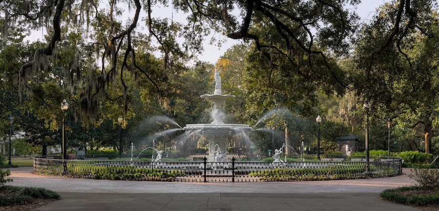 A fountain with water jets surrounded by a fence, set in a park with trees and a clear sky.