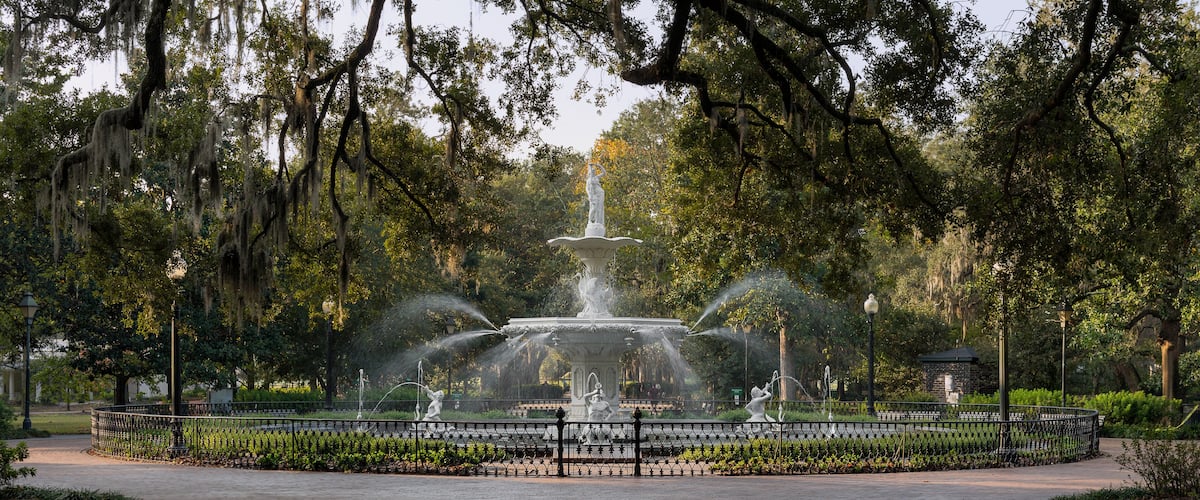 A fountain with water jets surrounded by a fence, set in a park with trees and a clear sky.
