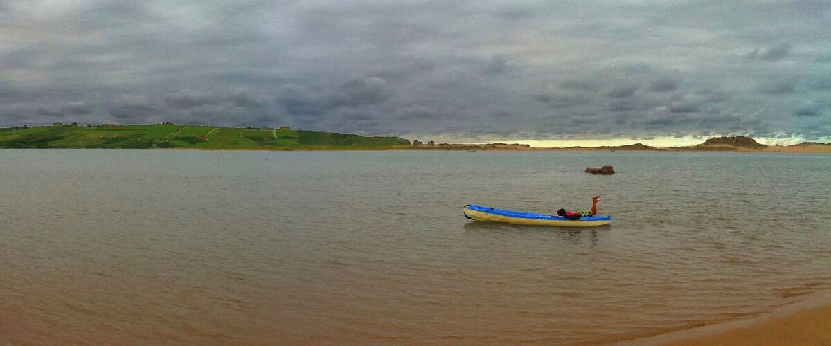 A calm body of water with a small blue and white kayak in the center, surrounded by a hilly landscape under a cloudy sky.