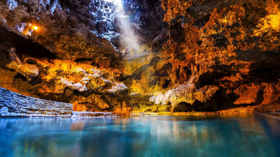 A cave with stalactites, a pool of water, and a light source illuminating the cave interior.