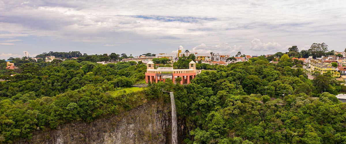 A waterfall cascading into a river surrounded by dense forest and a red building with a white dome in the background.