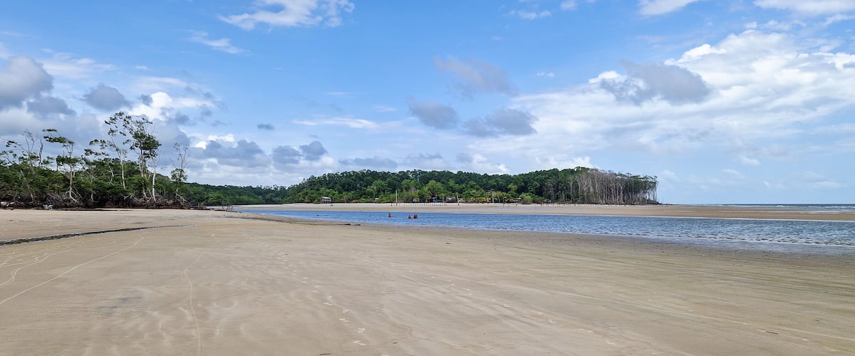 A wide sandy beach with gentle waves, a forested hill in the background, and a clear blue sky with scattered clouds.