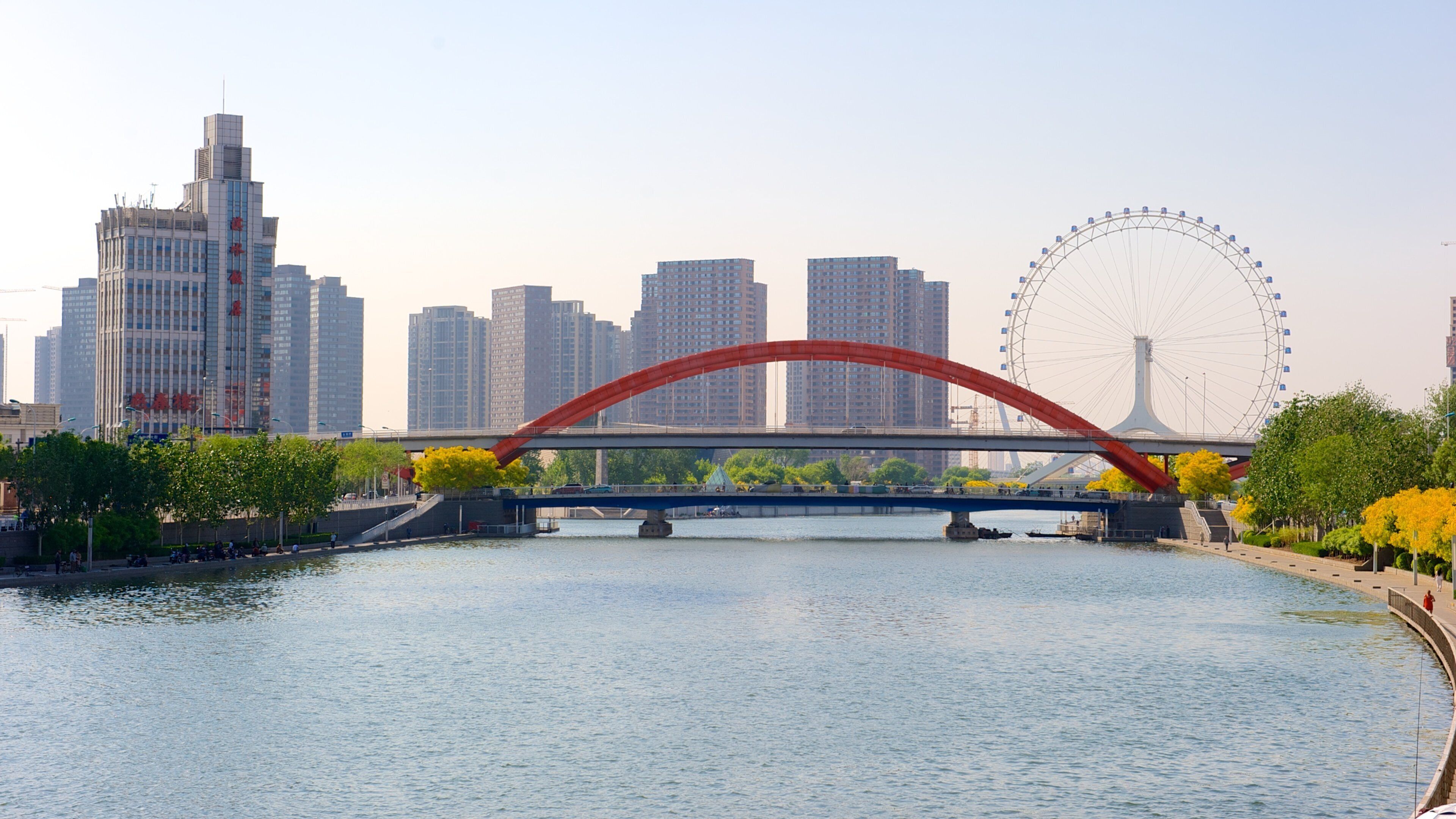 A river with a red bridge, a Ferris wheel, and modern buildings in the background.