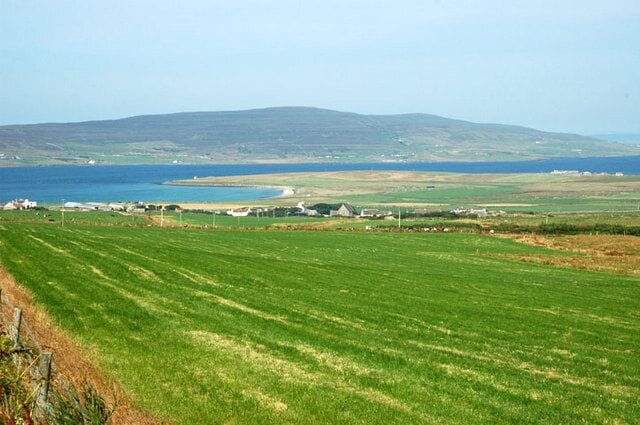 A coastal landscape with green fields, a body of water, and distant hills.