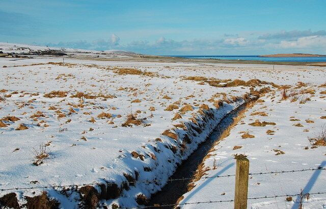 A snow-covered field with a drainage ditch and a wooden fence.