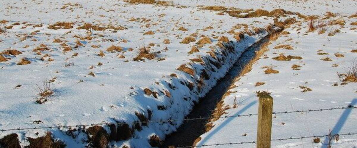 A snow-covered field with a drainage ditch and a wooden fence.
