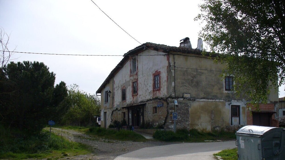 Ein verwittertes, zweistöckiges Haus mit einer unbefestigten Zufahrt, umgeben von viel Grün und einem Stromkasten.