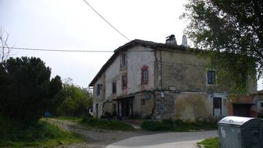 Uma casa antiga de dois andares com uma estrada de terra que a leva, cercada por vegetação e uma caixa de utilidades.