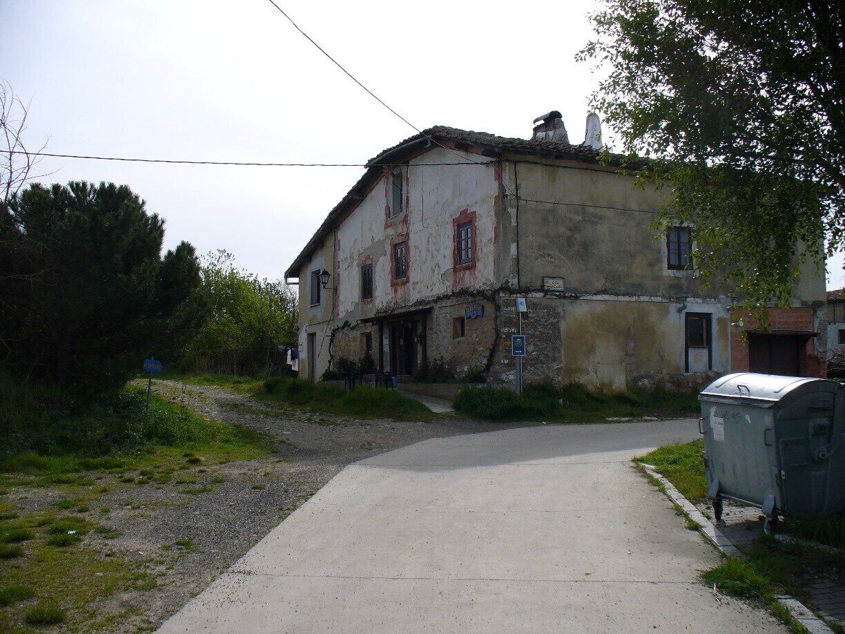 A weathered two-story house with a dirt road leading to it, surrounded by greenery and a utility box.