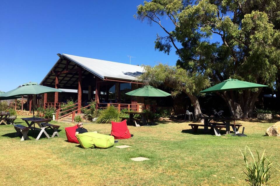 An outdoor seating area with picnic tables and green umbrellas, surrounded by trees and a building with a sloped roof.