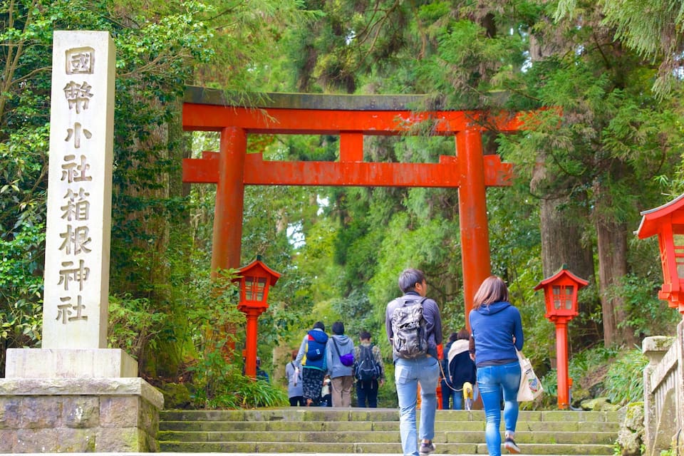 Um portão torii vermelho na entrada de uma área arborizada, com degraus de pedra que levam até ele.