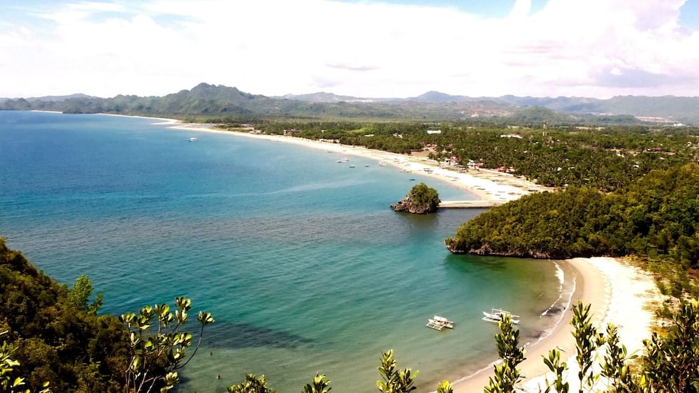 A coastal landscape with a beach, clear blue waters, and surrounding greenery.