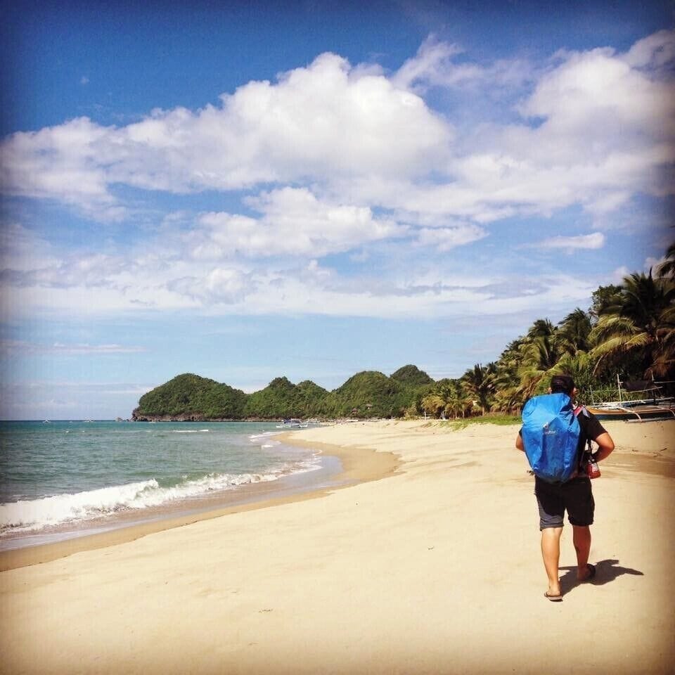 A beach with a person carrying a blue backpack, palm trees, and a small hill in the background.