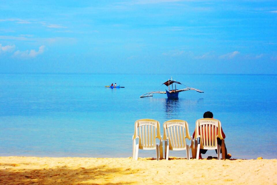 A beach with two chairs facing the sea, a boat with two people, and a seaplane flying low over the water.