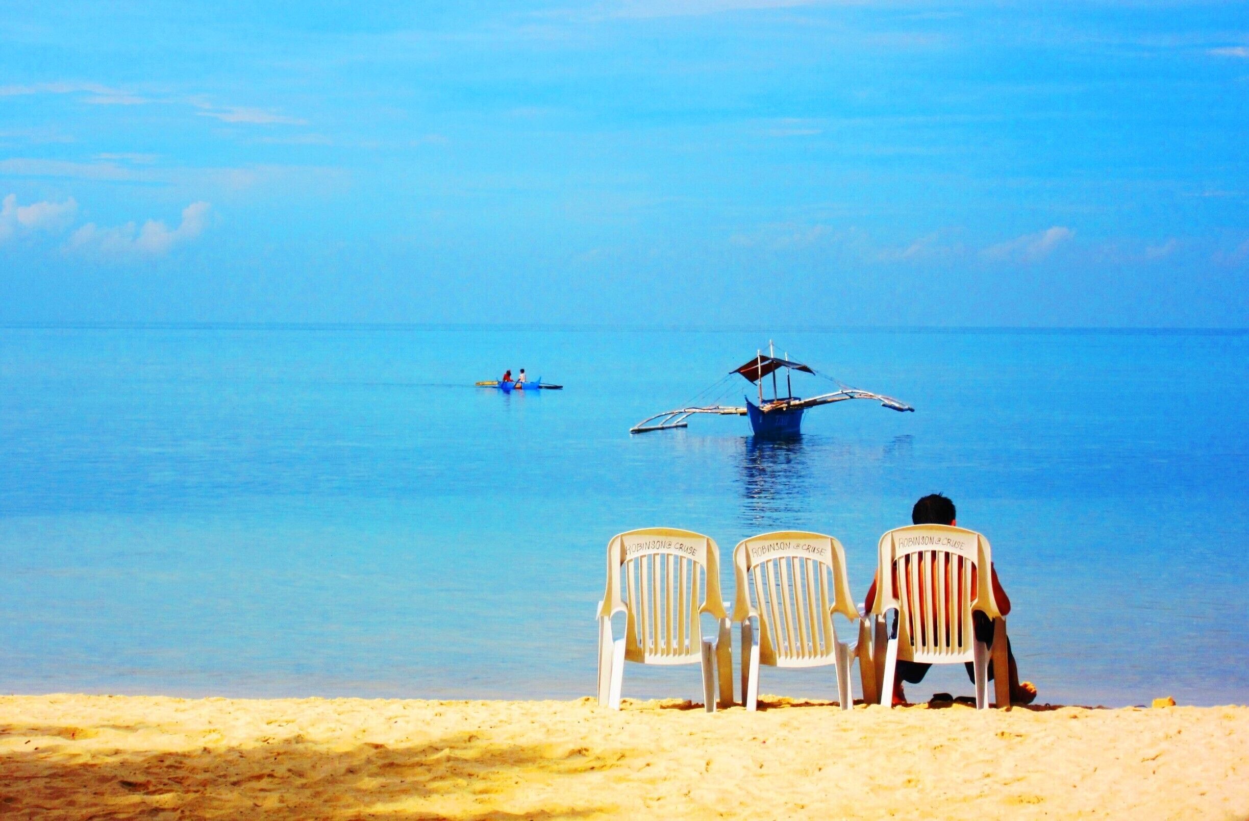 A beach with two chairs facing the sea, a boat with two people, and a seaplane flying low over the water.