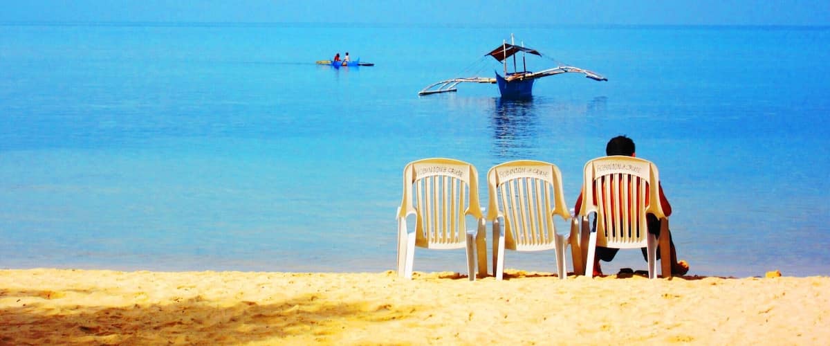 A beach with two chairs facing the sea, a boat with two people, and a seaplane flying low over the water.