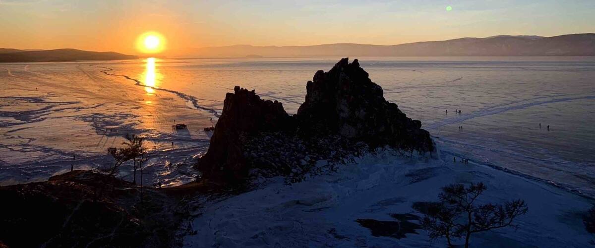 A frozen lake at sunset with a prominent rock formation in the foreground.
