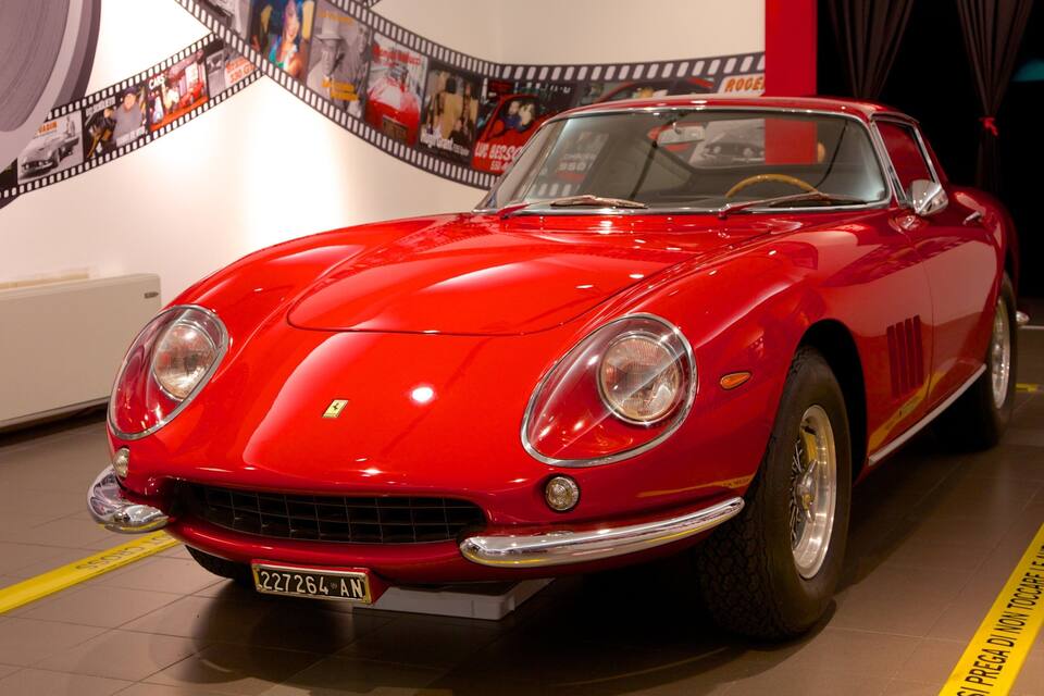 A red vintage car with a license plate, displayed indoors with a film strip backdrop.