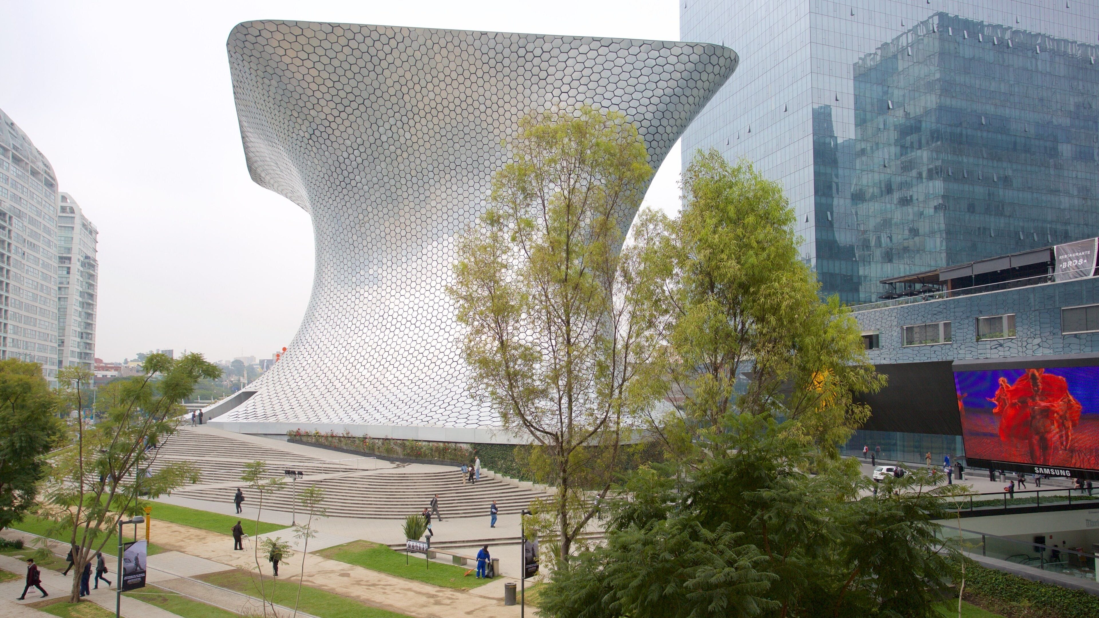 Uma grande escultura moderna numa praça pública, rodeada por edifícios e árvores.