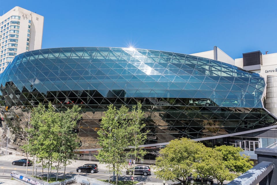 A modern glass building with a unique geometric design, surrounded by trees and other urban structures.