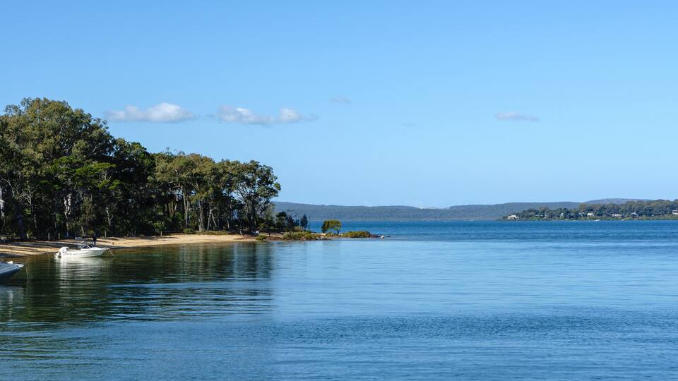 A calm lake with a boat docked on the left, surrounded by dense trees and a distant shoreline with buildings.