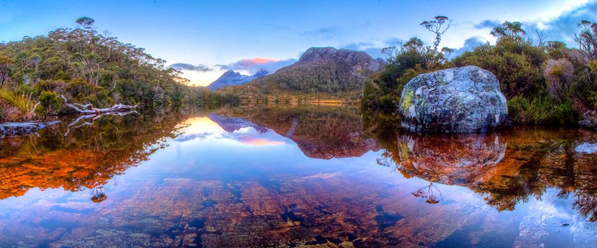 A calm lake reflecting trees and mountains, with a large rock in the foreground.