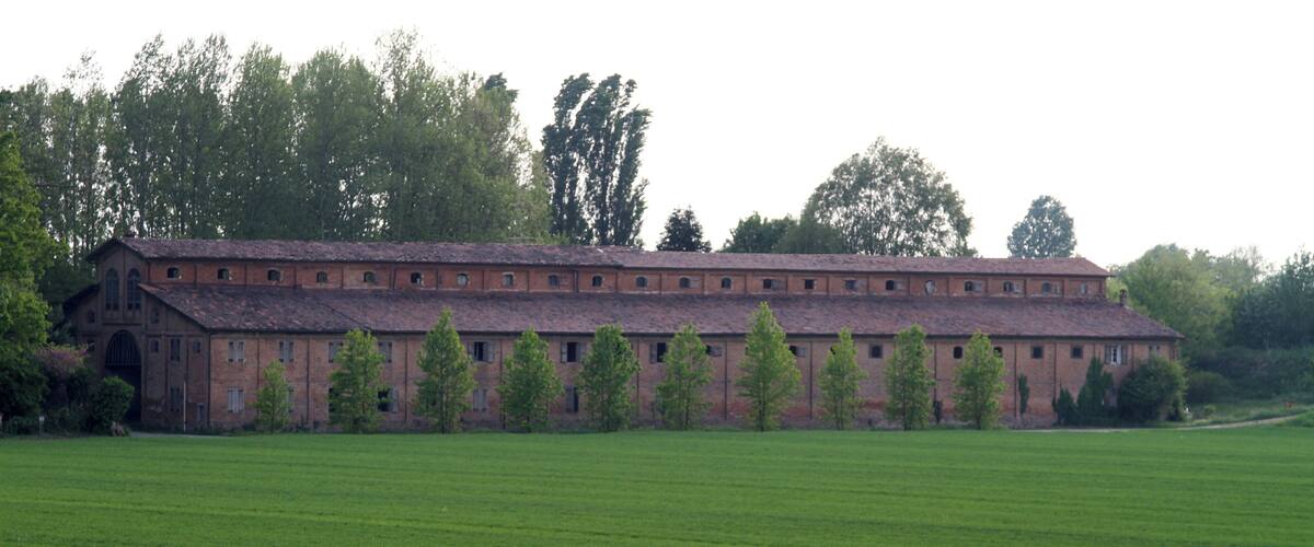 A large brick building with a long roof, surrounded by a green field and trees.