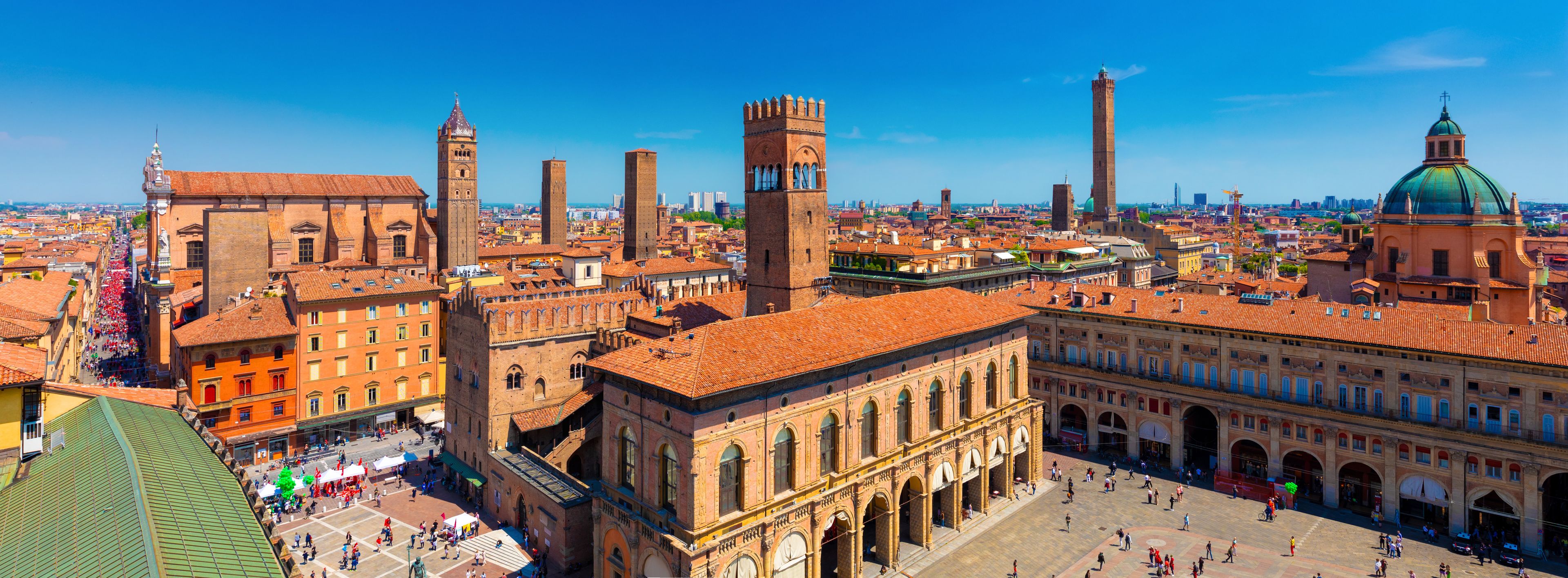 A historic cityscape with prominent red-tiled roofs, a large open square, and several tall towers and domes.