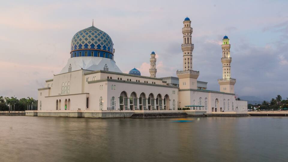 A mosque with a large blue dome and multiple minarets by a calm body of water.