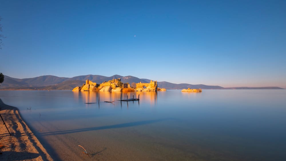 A calm lake with a rocky island featuring ruins, a clear sky, and distant mountains.