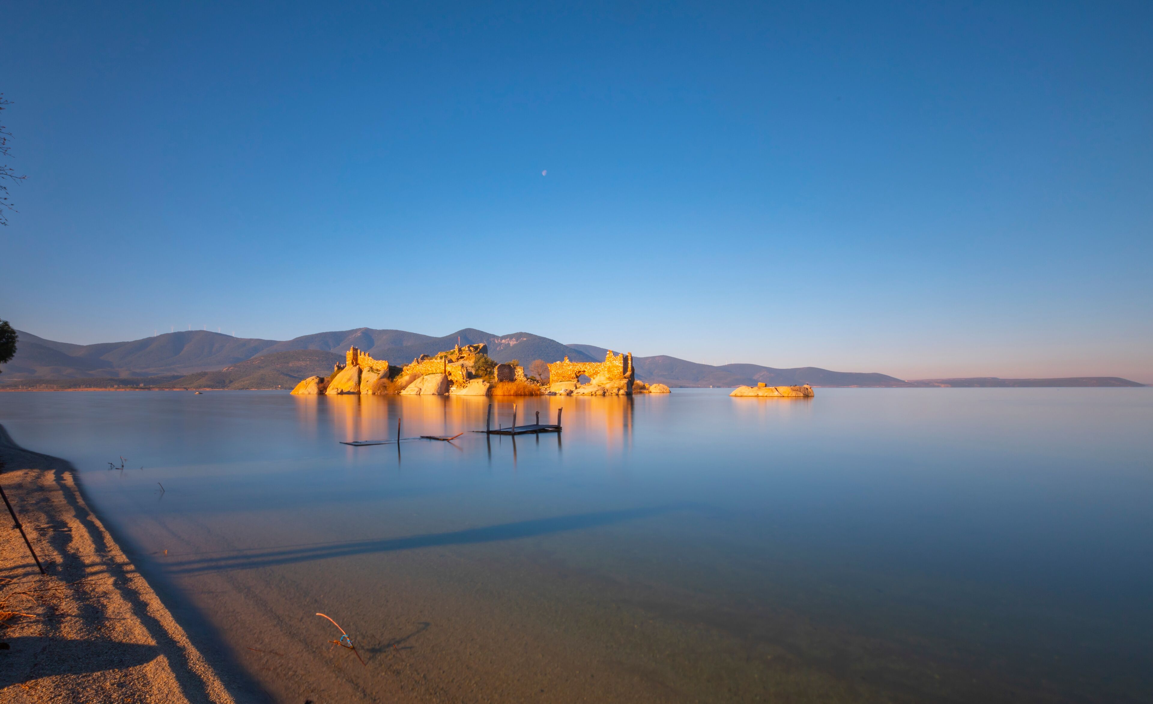 A calm lake with a rocky island featuring ruins, a clear sky, and distant mountains.