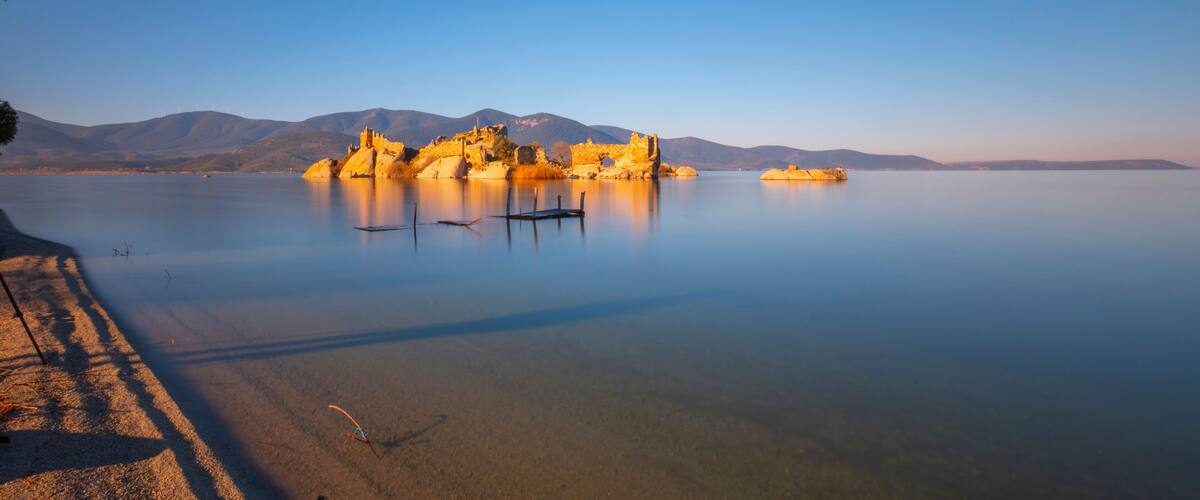 A calm lake with a rocky island featuring ruins, a clear sky, and distant mountains.