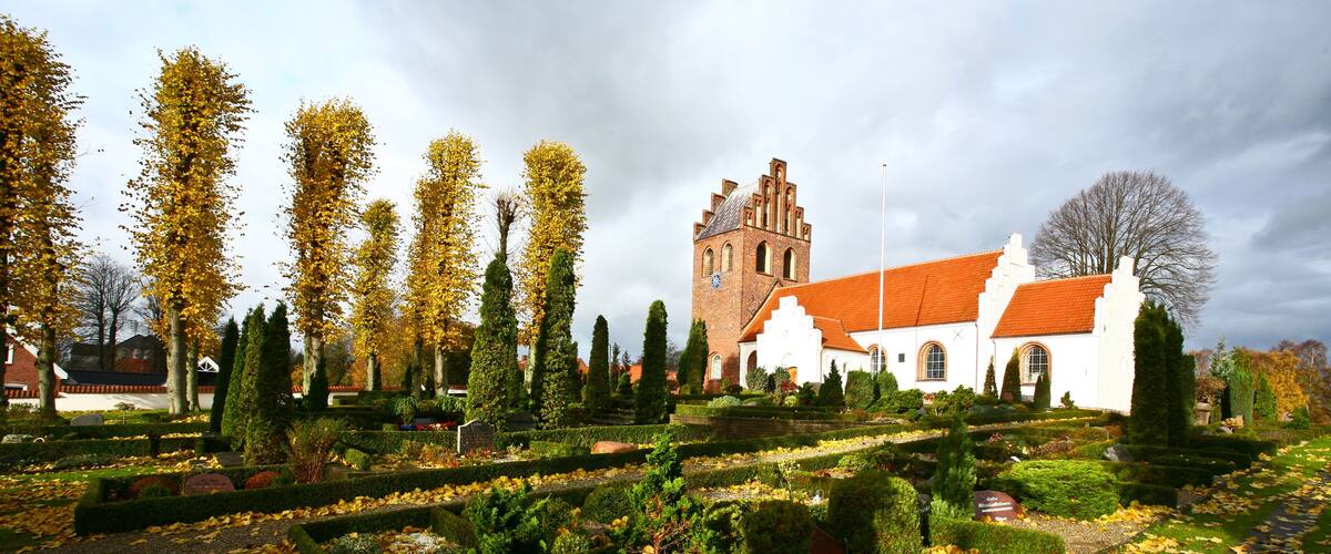 A historic church with a red roof, surrounded by a well-manicured garden with autumn leaves and tall trees.