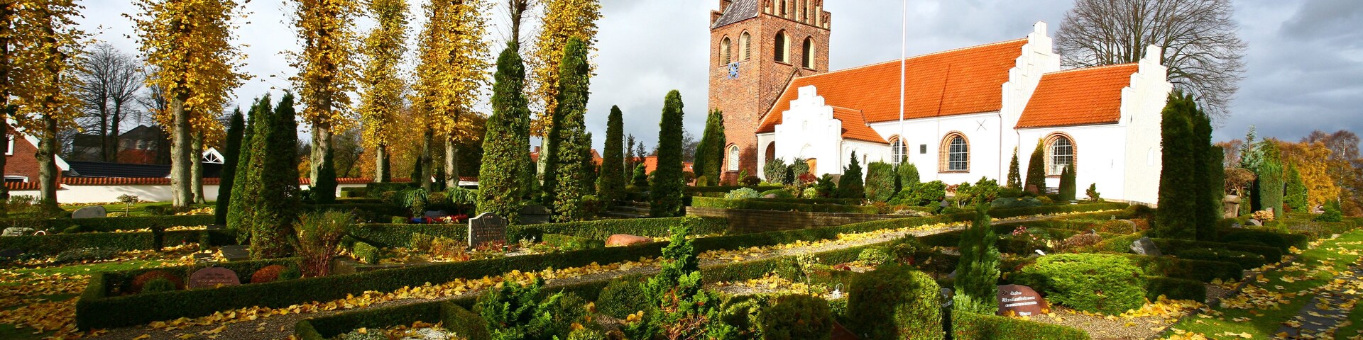 A historic church with a red roof, surrounded by a well-manicured garden with autumn leaves and tall trees.