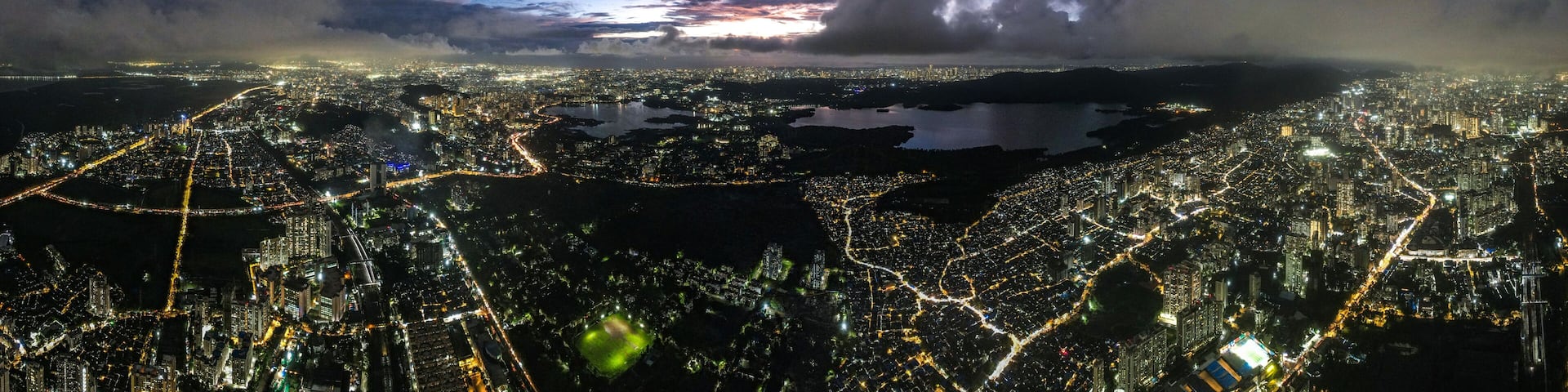 Aerial view of a sprawling cityscape with shimmering lights reflecting off Powai Lake under a dramatic twilight sky, Mumbai, Maharashtra, India.
