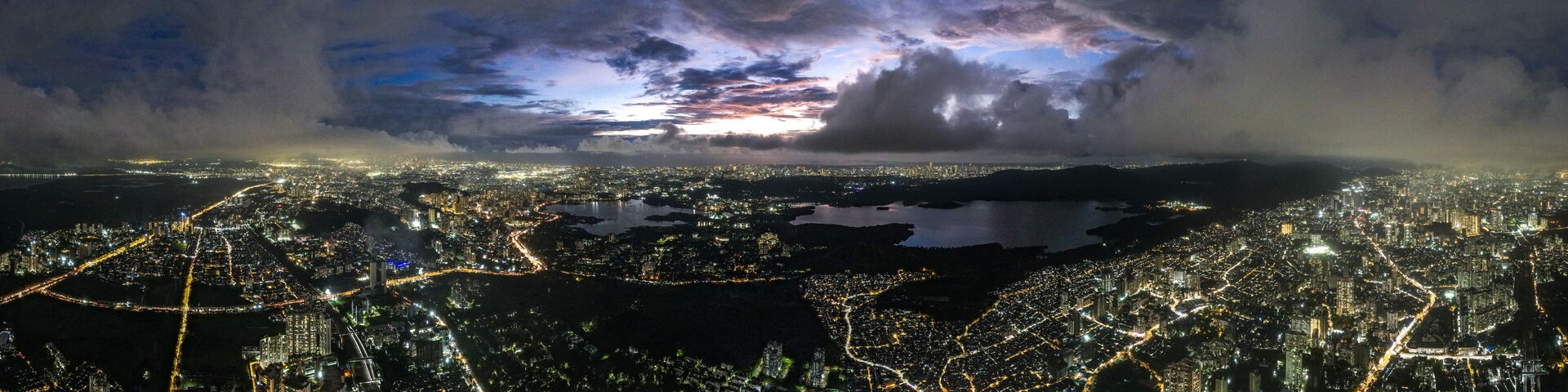 Aerial view of a sprawling cityscape with shimmering lights reflecting off Powai Lake under a dramatic twilight sky, Mumbai, Maharashtra, India.